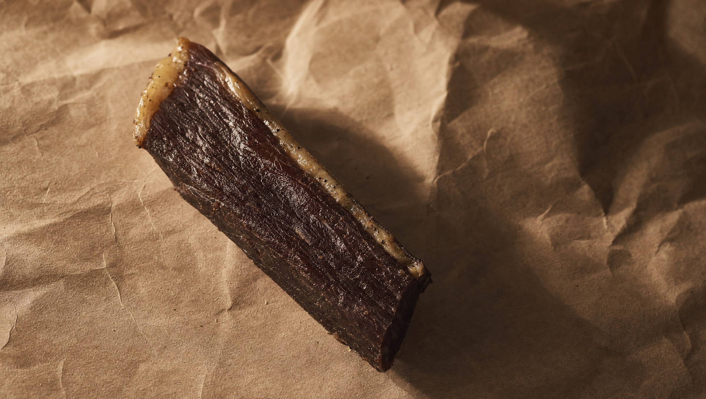 A single hand-cut biltong stick on warm brown butcher paper, hard raking side-light from the left, deep shadows. The family-recipe biltong made in the shop.